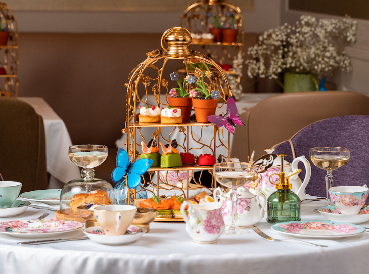 Themed afternoon tea London: a tiered afternoon tea stand in the form of a golden birdcage, decorated with colourful butterflies and flowers, filled with sandwiches, pastries and cakes, including one designed to look like a flower pot
