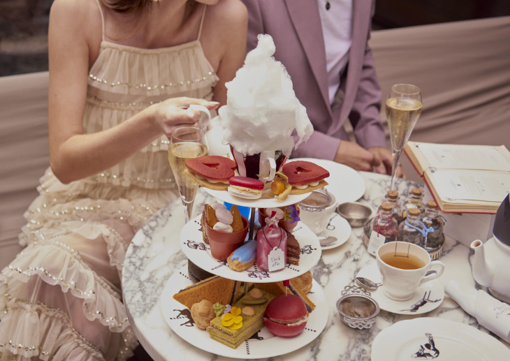 Themed afternoon tea London: a couple sitting at a table with a three-tiered stand, laden with goodies including red heart-shaped biscuits, a bottle labelled 'drink me' and candy floss frothing out of a mug.
